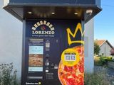 un distributeur automatique sur lequel est posée une pizza, devant un immeuble