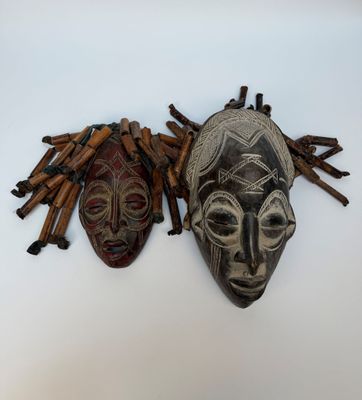 deux masques africains accrochés à un mur, à côté d'un tas de brindilles