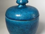 une image représentant un vase bleu muni d'un couvercle, posé sur une surface blanche