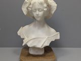 un buste blanc représentant une femme coiffée d'un chapeau, posé sur un socle en bois