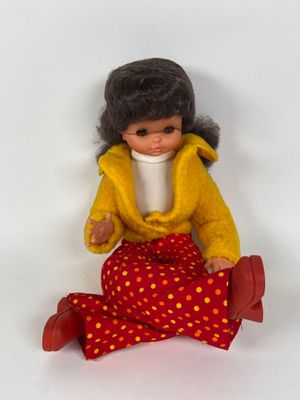 une petite poupée assise par terre, vêtue d'une veste jaune et d'un pantalon rouge