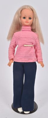 poupée aux cheveux blonds, vêtue d'un pantalon bleu et d'un pull rose, posée sur un socle noir