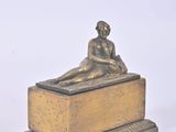 brass figurine of a woman sitting on a box on a white surface