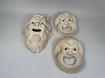 trois masques en céramique blanche représentant des visages, posés sur une surface blanche