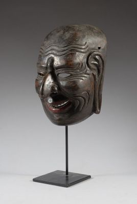 un masque en bois posé sur un socle, devant un fond gris