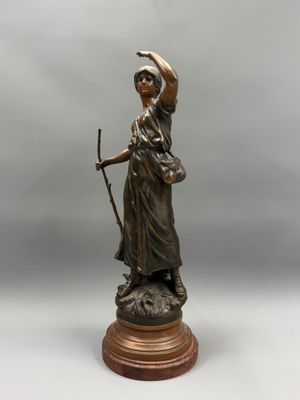 une statue antique en bronze représentant une femme tenant un bâton, posée sur un socle en bois