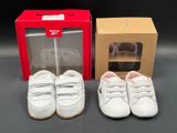two pairs of white baby shoes are next to a red box