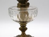 une lampe à huile ancienne en verre et laiton, posée sur un pied, sur fond blanc