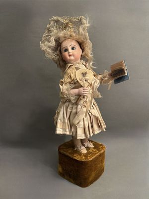une poupée ancienne tenant un livre et un chapeau, posée sur un bloc de bois