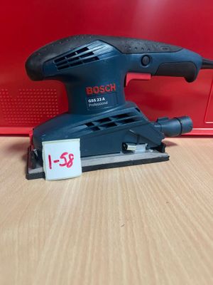 une ponceuse orbitale Bosch posée sur une table en bois, à côté d'une boîte rouge