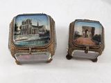pair of silver and glass boxes with paintings on them on a white surface