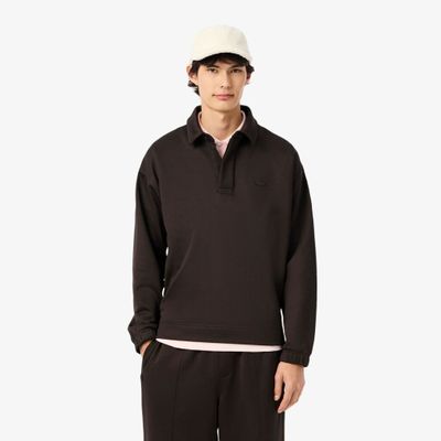 Un homme vêtu d'un polo noir et d'un pantalon se tient debout devant un fond blanc.