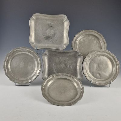 six plats et plateaux en métal argenté posés sur des supports, disposés sur une surface blanche
