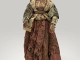 une vieille femme aux allures de poupée, vêtue d'une robe et d'un chapeau