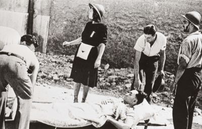 black and white photo of a group of people standing around a man laying on a bed