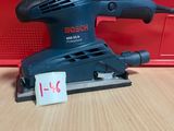 une ponceuse orbitale Bosch posée sur une table en bois, avec une boîte rouge en arrière-plan