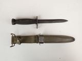 une paire de couteaux militaires posés sur une surface blanche, dont l'un se trouve au milieu des autres