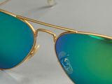lunettes de soleil à monture dorée et verres verts miroités, posées sur une surface blanche