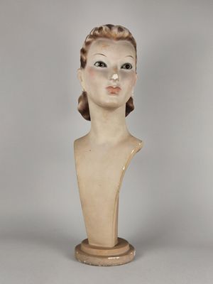 statue représentant la tête d'une femme, posée sur un socle en bois, sur fond gris