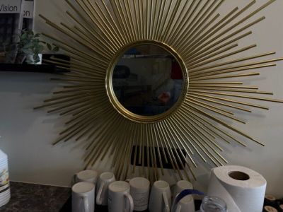 une photo d'un miroir doré en forme de soleil accroché au mur, au-dessus de tasses à café