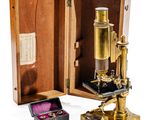 un microscope en or posé dans un coffret en bois, à côté d'une bague rose