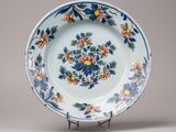 assiette décorée de fleurs et de feuilles, posée sur un socle, sur fond gris