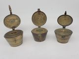 trois récipients en laiton avec leurs couvercles, posés sur une surface blanche