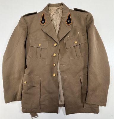 La veste est de couleur beige clair et comporte des boutons dorés au niveau du col.