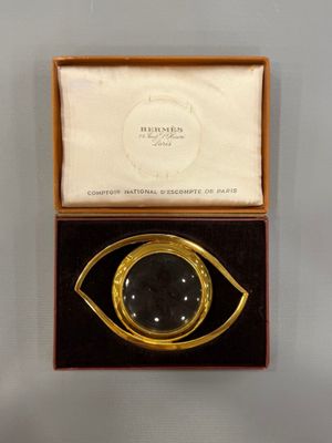 une photo d'une broche en forme d'œil, dorée et noire, dans une boîte