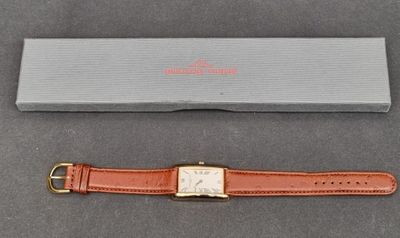 une montre marron avec une boucle dorée et un écrin posés sur un fond gris