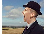 un homme portant un chapeau et un costume, la bouche grande ouverte, devant un avion