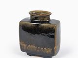 un vase noir avec un couvercle marron posé sur une surface blanche