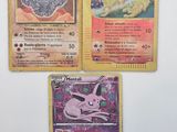 Trois cartes Pokémon sont posées sur une surface blanche ; l'une est rose et l'autre est jaune.