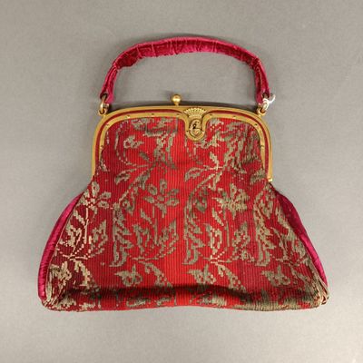 une image représentant un sac à main rouge muni d'une poignée dorée, posé sur une surface grise