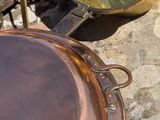 une grande casserole en cuivre posée par terre à côté d'une marmite et d'une poêle