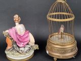 two figurines of a woman sitting on a bench next to a birdcage