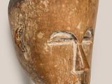 une photo représentant un masque en bois posé sur un socle devant un mur blanc