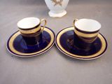 two cups and saucers are sitting on a table with a vase in the background