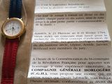 une montre posée sur une feuille de papier, à côté d'une autre feuille de papier à laquelle est attachée une feuille de papier