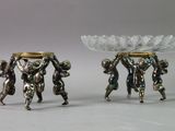 two silver figurines that are holding a bowl on their heads