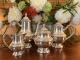 trois théières en argent posées sur une table, à côté d'un vase de fleurs
