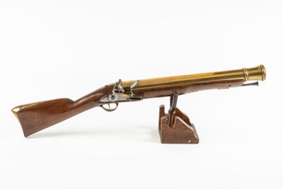 un pistolet ancien plaqué laiton, posé sur un socle en bois, sur fond blanc
