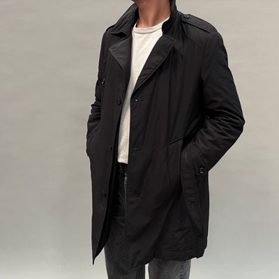 un homme vêtu d'un manteau noir et d'un jean, debout, les mains dans les poches