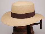 un chapeau de paille orné d'un ruban marron, posé sur un porte-chapeaux en bois