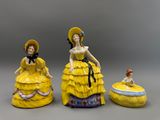 trois figurines en porcelaine représentant des femmes vêtues de robes jaunes et coiffées de chapeaux