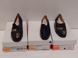 trois paires de chaussures noires posées sur des cartons, sur une surface blanche