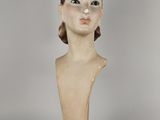 statue représentant la tête d'une femme, posée sur un socle en bois, sur fond gris