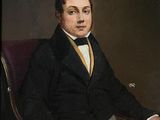 portrait of a man in a black jacket and yellow tie sitting in a red chair