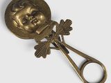 cuillère plaquée or sur laquelle est représentée une tête de poupée, avec une paire de ciseaux devant