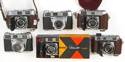 six old cameras are lined up on a white surface with a red apple